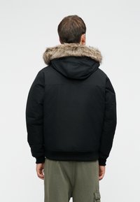 Μαύρο μπουφάν τύπου πούφτερ με κουκούλα επενδυμένη με γούνα, διαθέτοντας λεία υφή, ριγωτούς καρπούς και άνετη εφαρμογή. Απλός σχεδιασμός χωρίς ορατά λογότυπα.