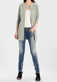 Svetle zelený texturovaný kardigan s kulatým výstřihem, otevřeným předním dílem a dlouhými rukávy. Kombinován s roztrhanými modrými skinny džínami a černými lodičkami na otevřenou špičku.