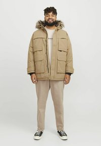 Parka beige con capucha y ribete de piel, cuatro bolsillos frontales y cierre con cremallera. Lleva puesta una camisa beige clara y combina con pantalones claros y zapatillas.