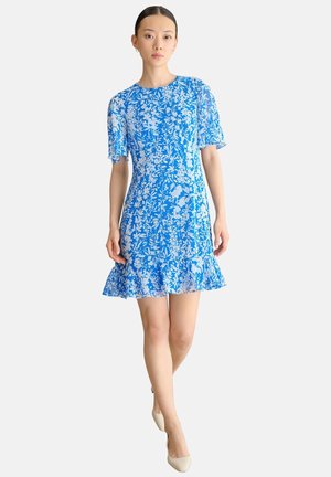 Mujer que lleva un vestido de manga corta con estampado floral azul y blanco y un dobladillo con volantes, de pie con una pierna ligeramente adelantada sobre un fondo blanco.
