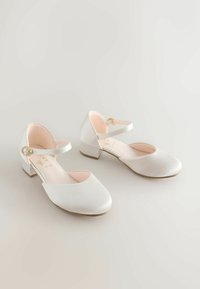 Valkoiset nahkaiset korolliset kengät pyöreällä kärjellä ja nilkkaremmin kanssa. Varustettu pienellä kultaisella soljella ja materiaalissa on teksturoitu pinta.