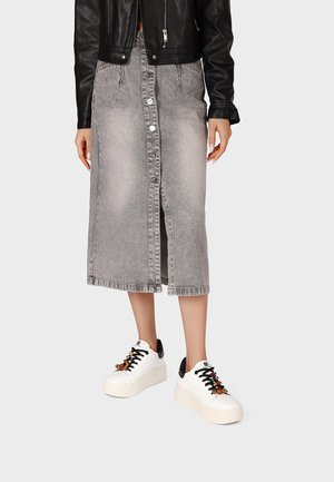 Persona che indossa una gonna midi di jeans grigia con bottoni, giacca di pelle nera e sneakers bianche con plateau, lacci neri e perline decorative.