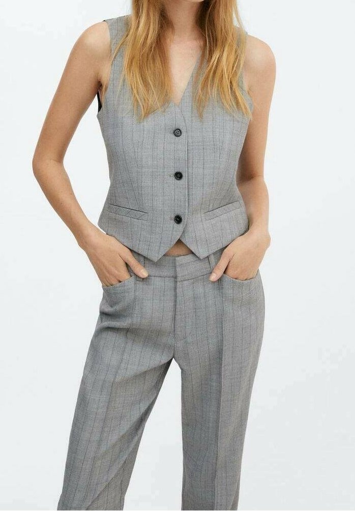 Femme portant un gilet sans manches gris à fines rayures avec des boutons noirs et un pantalon taille haute assorti, les mains dans les poches du pantalon.