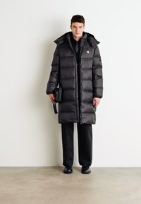 Schwarze Daunenjacke mit Kapuze, ausgestattet mit einem Reißverschluss und sichtbaren Nähten. Über schwarzer Kleidung getragen, vor einem schlichten Hintergrund.