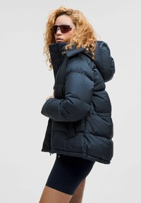 Doudoune de couleur bleu marine foncé, avec un col haut et une capuche, un design matelassé et des poches avant. Portée par-dessus un short noir.