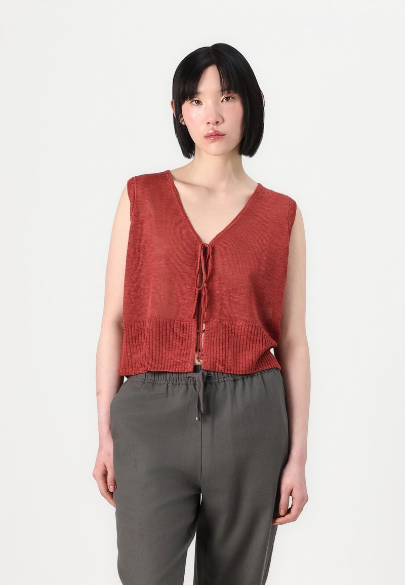 Rustfarget cropped vest med V-hals, ribbestrikket kant og bindebånd foran, paired med løse grå bukser. Ensfarget bakgrunn.