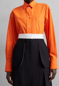 Oranssi puuvillapaita, jossa on napitus edessä ja rintatasku, yhdistetty mustaan hameeseen, jossa on valkoisia yksityiskohtia ja laskostettu muotoilu.
