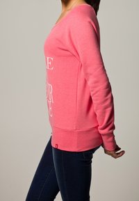 Roze sweatshirt met lange mouwen en een brede halslijn, voorzien van witte tekst op de voorkant. Zachte stof, losvallende pasvorm en geribbeldezoom.