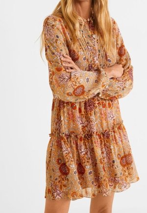 Vrouw met lang blond haar, gekleed in een losse, lange mouw jurk met bloemenprint in warme herfstkleuren, met gekruiste armen.