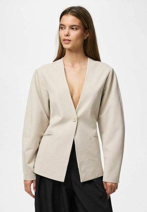Femme portant un blazer beige à un bouton avec un décolleté profond et un pantalon noir à jambes larges, debout devant un fond blanc uni.