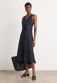 Mørkeblå ermeløs kjole med dyp V-hals, med en samlet midje og flytende skjørt. Kombinert med svarte slip-on mules og en tote-veske.