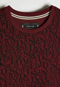 Bordowa koszulka z okrągłym dekoltem, z powtarzającym się czarnym wzorem tekstu "DIVERSE" oraz czarną metką z białym napisem "diverse" wewnątrz kołnierzyka.