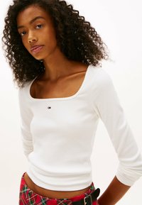 Mujer joven con cabello rizado que lleva una blusa blanca de manga larga con cuello cuadrado y una falda de cuadros rojos con un cinturón negro.