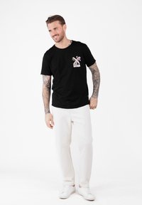 Hombre con tatuajes en los brazos vistiendo una camiseta negra con un diseño de molino de viento floral, pantalones blancos y zapatillas blancas frente a un fondo blanco.