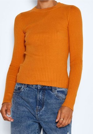 Person iført en tætsiddende, ribbet, orange langærmet sweater kombineret med højtaljede, falmede blå jeans og en guldring på højre hånd.