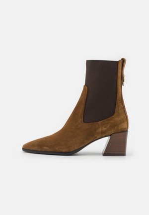 Bottine en daim marron avec bout pointu, panneaux latéraux élastiques et talon empilé. Dispose d'une tige en tissu marron et d'un accent argenté.