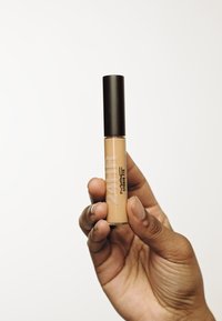 Concealer-Röhre mit transparentem beigen Gehäuse und einer dunklen, mattschwarzen Kappe. Mit dem Text "24HR" und "Studio Fix" in fetter Schrift.
