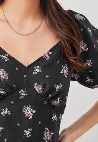Femme portant une robe noire à imprimé floral rose, manches bouffantes, encolure en V, et un délicat collier en perles dorées.