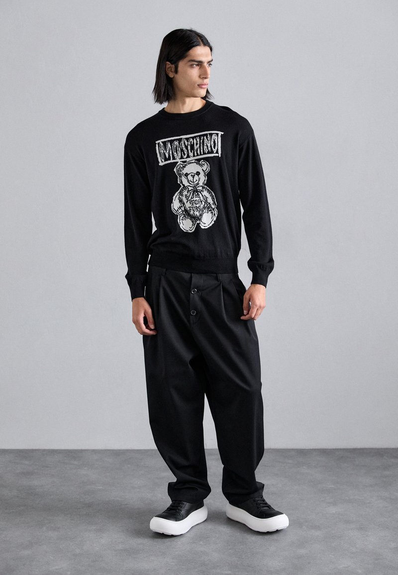Zwarte trui met een graphic van een beer met de tekst "Moschino" erboven. Gecombineerd met ruime zwarte broeken en chunky zwart-witte schoenen.