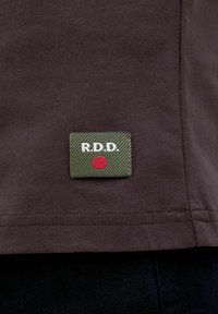 Temno rjava majica s tkanim zelenim obližem, ki prikazuje "R.D.D." v belem besedilu in rdečo piko spodaj, zaključenim z zavihkom.