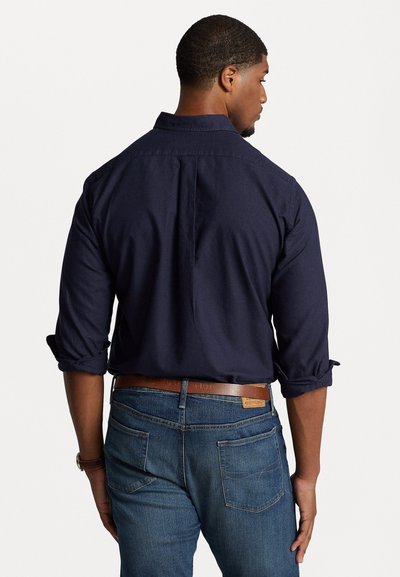 Chemise à manches roulées bleu marine avec boutons, présentant un empiècement au dos et une texture lisse, associée à un jean en denim et à une ceinture en cuir marron.