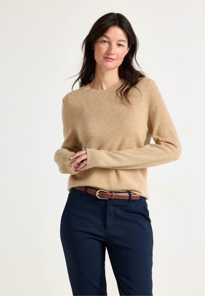 Vrouw met donker haar, gekleed in een beige trui met lange mouwen, marineblauwe broek en een bruine riem, staand tegen een effen lichte achtergrond.
