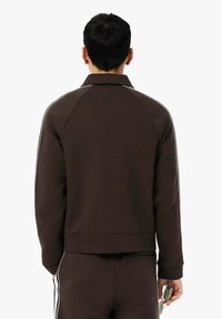 Chaqueta marrón con cremallera y mangas raglán, con franjas blancas a los lados, textura suave y cintura ajustada. Combinada con pantalones a juego.