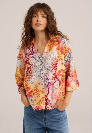 Femeie cu păr castaniu ondulat, purtând o cămașă colorată cu imprimeu floral și blugi albaștri, stând cu mâinile în buzunare, pe fundal simplu.