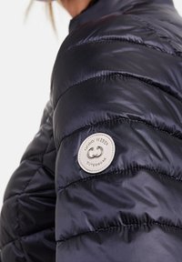 Marinblå puffjacka med kviltadtextur. Har en rund logotyp-patch på ärmen. Materialet verkar slätt och blanka.