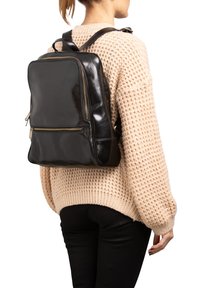 Zaino in pelle nera con finitura liscia, forma rettangolare, due scomparti con zip e spallacci regolabili, indossato sopra un maglione a maglia beige.