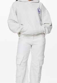 Sudadera de gran tamaño en gris claro, fabricada en un tejido suave, con un bolsillo frontal y la impresión "NO MOLESTAR". Combinada con pantalones cargo blancos con bolsillos laterales.