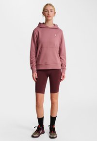 Kapuzensweatshirt in staubigem Rosa, mit einer Vordertasche und gerippten Bündchen, kombiniert mit burgunderfarbenen Radshorts und mehrfarbigen Sportschuhen.