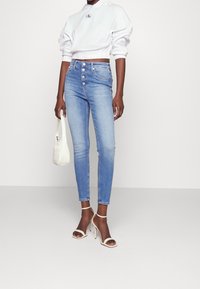 Obcisłe dżinsy w jasnoniebieskim kolorze, z wysokim stanem i zapięciem na guziki. Połączone z białą krótka bluzą z kapturem i beżowymi szpilkami na kostce z paskiem.