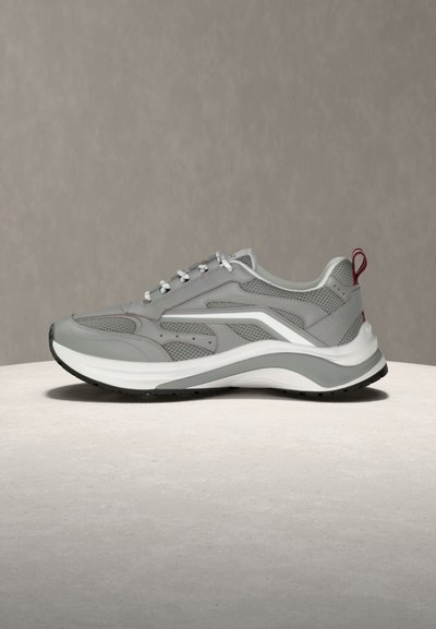 Γκρι αθλητικό παπούτσι με πάνελ από δίχτυ και δέρμα, λευκά κορδόνια, παχύ μαλακό πέλμα και κόκκινη θηλιά στο τακούνι, τοποθετημένο σε φωτεινή επιφάνεια.