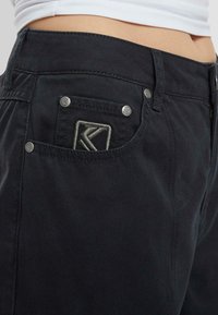 Czarne dżinsy o dopasowanej talii, z wyszywanym logo na przedniej kieszeni i metalowymi elementami guzika. Gładka faktura.