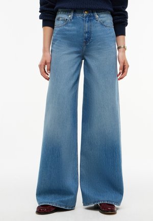 Persona con vaqueros azules de tiro alto y pata ancha, zapatos rojos y un suéter azul oscuro, de pie contra un fondo blanco.