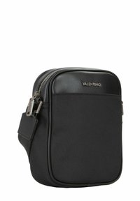 Sac bandoulière en tissu noir avec un dessus en cuir lisse, fermeture éclair, et forme rectangulaire. Comprend une petite étiquette près de la fermeture éclair.