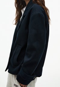 Kobieta nosząca czarną marynarkę z mieszanki wełny na białej bluzce oraz jasnoszarych dresach z logo na udzie, stojąca na białym tle.