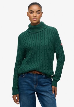 Grøn kabelstrikket sweater med høj krave, med ribbede manchetter og kant, båret med blå denimjeans og et vævet bælte. Rød insignie detalje på ærmet.
