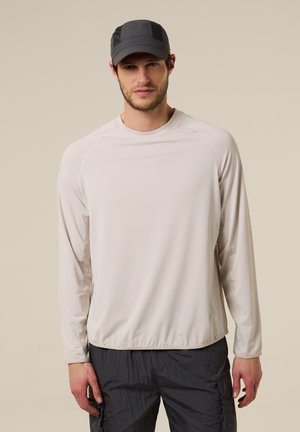 Hombre con camisa de manga larga beige claro, pantalones cargo negros y gorra de béisbol gris oscuro, de pie contra un fondo beige liso.