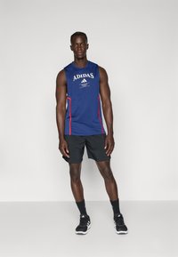 Sininen hihaton Adidas-toppia, jossa on valkoinen logo, punaiset sivuvihjeet ja mustat shortsit. Mustat urheilukengät ja sukat täydentävät asun.