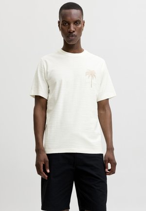 Man met een wit T-shirt met een klein beige palmboommotief op de borst en een zwarte korte broek, staand tegen een effen lichte achtergrond.