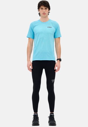 Young man standing, wearing light blue short-sleeve athletic shirt, black compression leggings, black running shoes, and a black wristwatch.