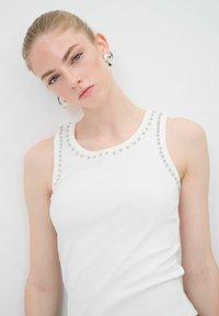 Top bianco senza maniche realizzato in tessuto liscio, con borchie argentate lungo il colletto. La modella indossa orecchini a cerchio semplici e un trucco naturale.