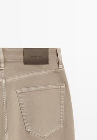 Vue arrière d'un jean en denim beige montrant la poche arrière droite et l'étiquette de marque en cuir marron "Massimo Dutti" sur la ceinture.
