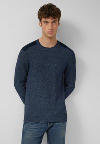 Navy gestrickter Pullover mit strukturiertem Stoff, rundem Halsausschnitt und dunklen Schulterakzenten. Getragen mit blauen Jeans, die ein Logo-Tag am Saum haben.