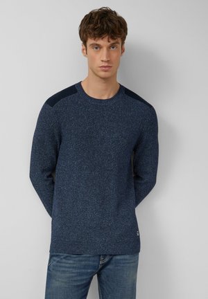 Gebreide marineblauwe trui met gestructureerde stof, ronde halslijn en donkere schouderaccenten. Draagt met blauwe jeans, voorzien van een logotag aan de zoom.