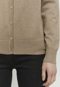 Beige cardigan with a fine knit texture, featuring a button-down front, ribbed cuffs, and hem, paired with dark jeans.