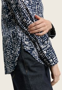 Dunkelblaues gemustertes Hemd mit weißen abstrakten Punkten, ausgestattet mit einem Knopfverschluss an den Bündchen und einem abgerundeten Saum, getragen mit dunklen Jeans.