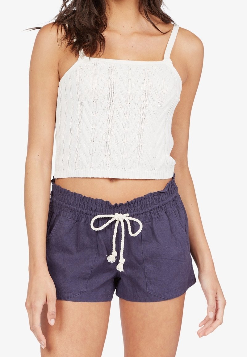 Femme portant un crop top blanc en maille et un short bleu marine avec une taille élastique et un cordon blanc noué en nœud.